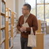 図書館の本棚の前で、借りた本と布の鞄を持つ一人暮らしの男性