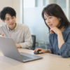 会議室で新しい受注システムを操作する48歳の女性と、説明する若手の同僚