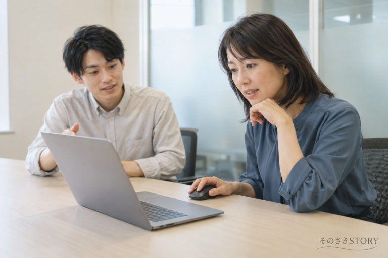 会議室で新しい受注システムを操作する48歳の女性と、説明する若手の同僚