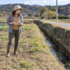 相続した畑の水路のそばで、土地の形を紙に描きながら周囲を確かめる54歳の智佳を描いたイメージ