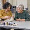 手芸の会で補聴器をつけ、隣の人が見せる縫い目を確かめながら話す76歳の鮎子のイメージ