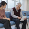 病院の待合室で、自分の質問メモを持つ78歳の父と、隣で話を聞く50歳の娘・沙知を描いたイメージ